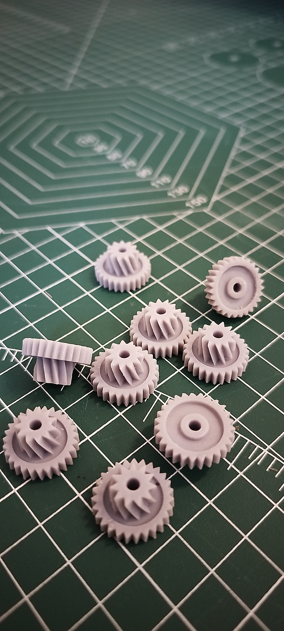 Szare, wydrukowane w 3D koła zębate na zielonej macie z siatką, ułożone w nieładzie. Dwa koła ustawione jedno na drugim. Widoczne detale druku 3D.