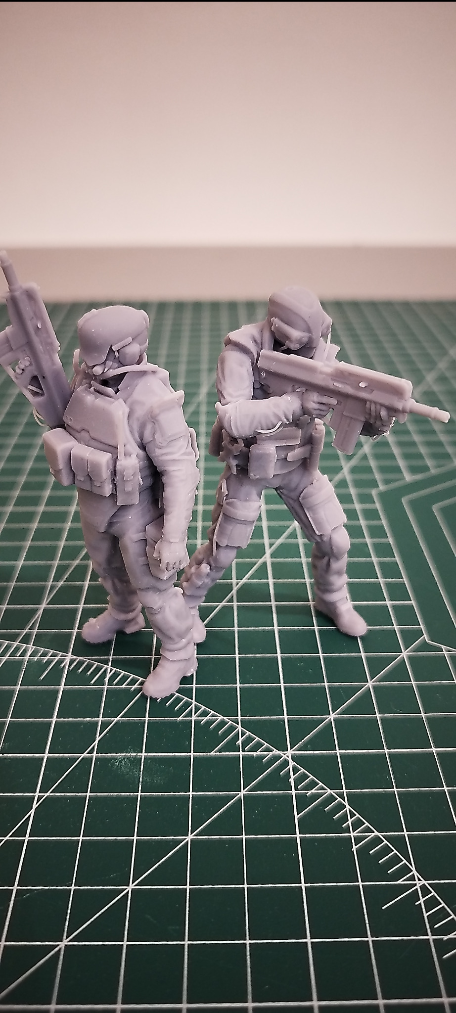 Dwie szare figurki żołnierzy z bronią, wydrukowane w 3D, stojące na zielonej macie z podziałką. Widoczne detale wyposażenia i broni.