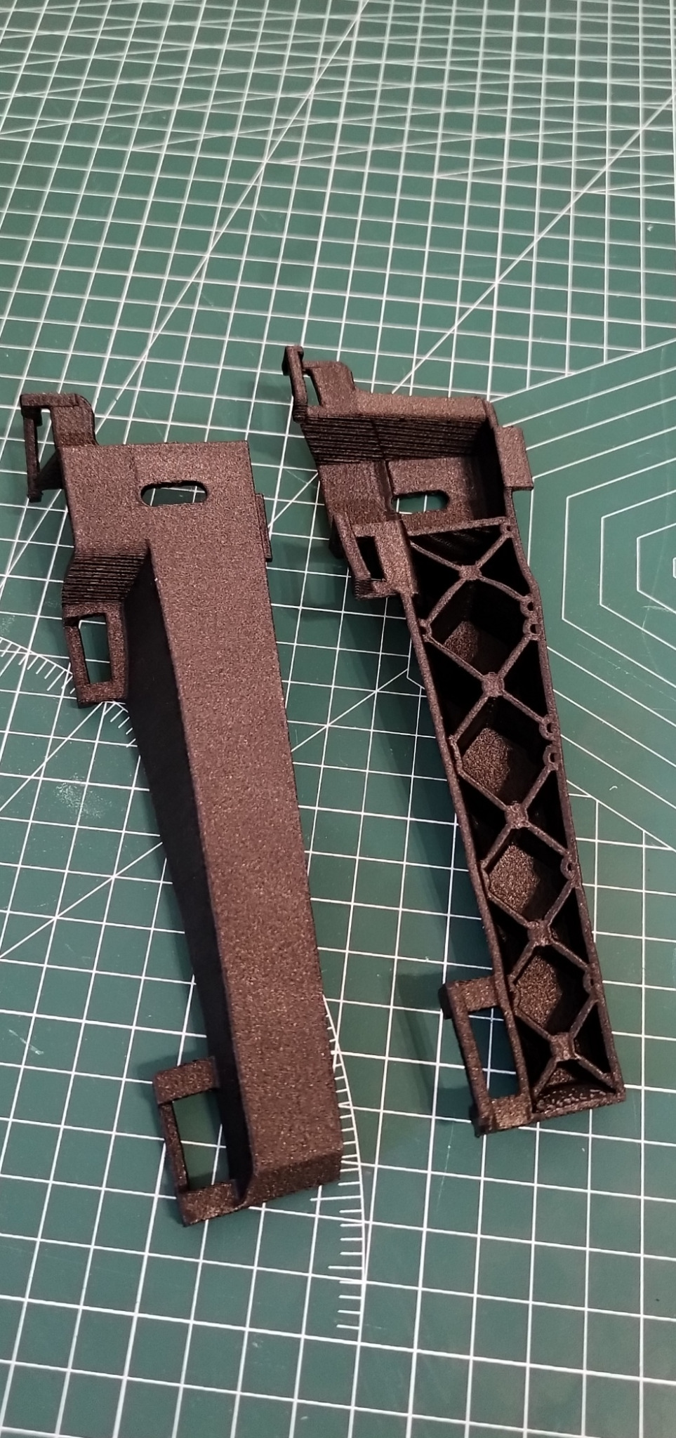 Dwa brązowe, wydrukowane elementy z tworzywa sztucznego na zielonej macie. Jeden element pełny, drugi z ażurową konstrukcją w kształcie rombów.