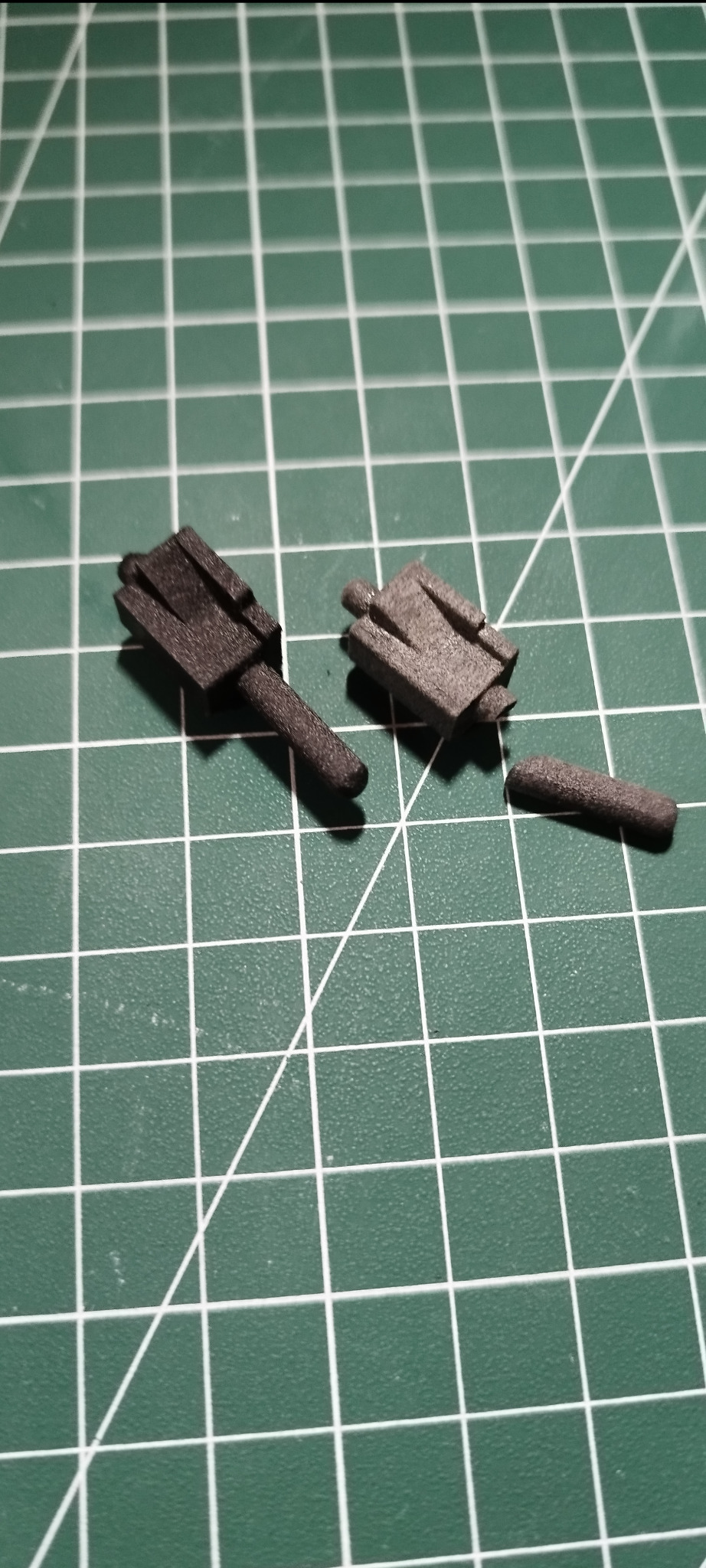 Dwa ciemne, matowe elementy z drukarki 3D leżą na zielonej macie z siatką. Jeden element jest uszkodzony, z odłamanym fragmentem. Widoczne ślady druku warstwowego.