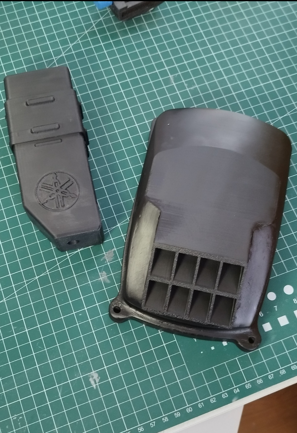 Czarne, matowe elementy z tworzywa sztucznego, prawdopodobnie obudowy, wydrukowane w 3D, leżą na zielonej macie. Jeden element z logo Yamaha.