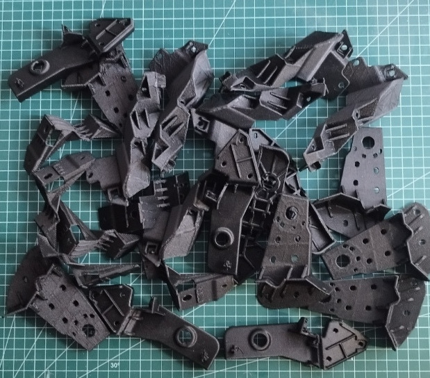 Czarne, matowe elementy z tworzywa sztucznego, wydrukowane w 3D, rozłożone na zielonej macie pomiarowej. Widoczne otwory i struktura warstw wydruku.