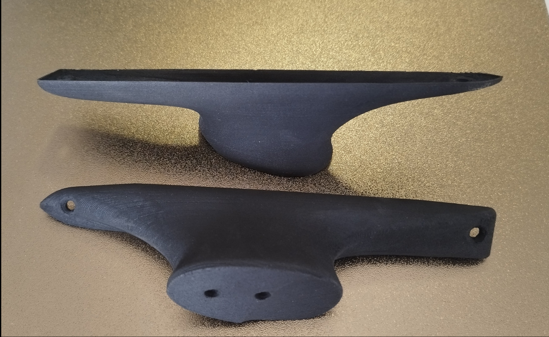 Dwa czarne, matowe elementy z tworzywa sztucznego, wydrukowane w 3D, leżą na złotej powierzchni. Widoczne warstwy druku i otwory montażowe.