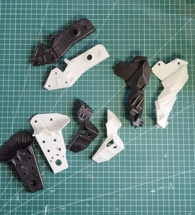 Różne, czarno-białe elementy z tworzywa sztucznego, prawdopodobnie prototypy lub części zamienne, ułożone na zielonej macie z podziałką. Widoczne otwory i mocowania.