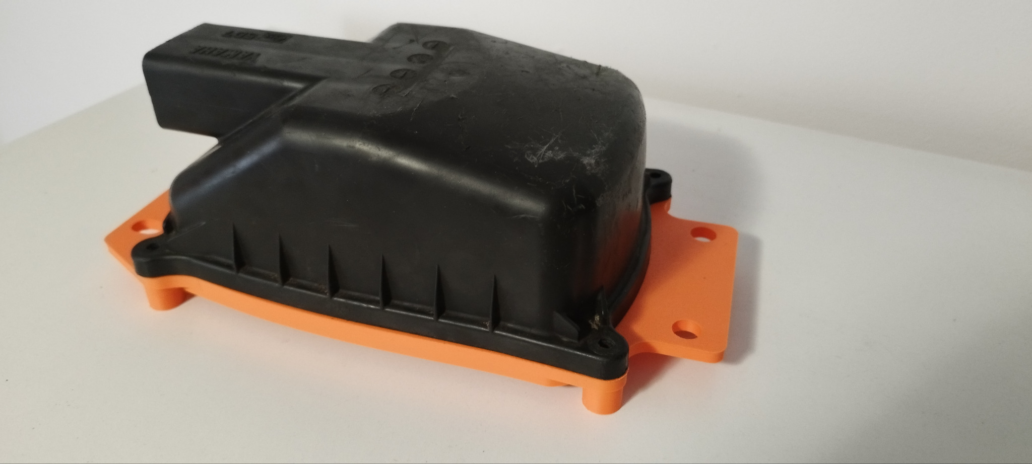 Czarna obudowa filtra z pomarańczową podstawą, wydrukowana w 3D, leży na białym tle. Widoczne detale druku i otwory montażowe.