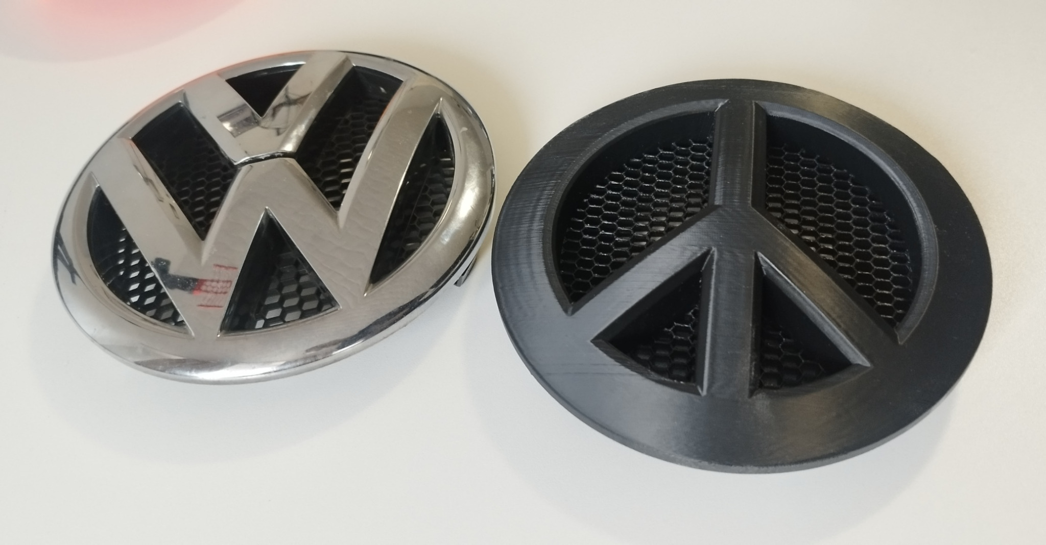 Porównanie: chromowany znaczek VW i czarny, drukowany 3D zamiennik z symbolem pacyfistycznym. Widoczna struktura druku 3D i siatka w tle.