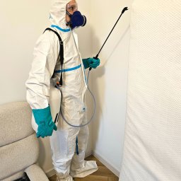 Sanito Paweł Stąsiek - Osoba w kombinezonie ochronnym i masce spryskuje materac. Widoczny aplikator, butla na plecach i fragment sofy. Dezynfekcja materaca w pomieszczeniu.