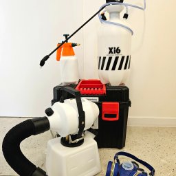 Sanito Paweł Stąsiek - Profesjonalny sprzęt do dezynfekcji: zamgławiacz, opryskiwacze ciśnieniowe i maska ochronna na tle białej ściany i podłogi z płytek.