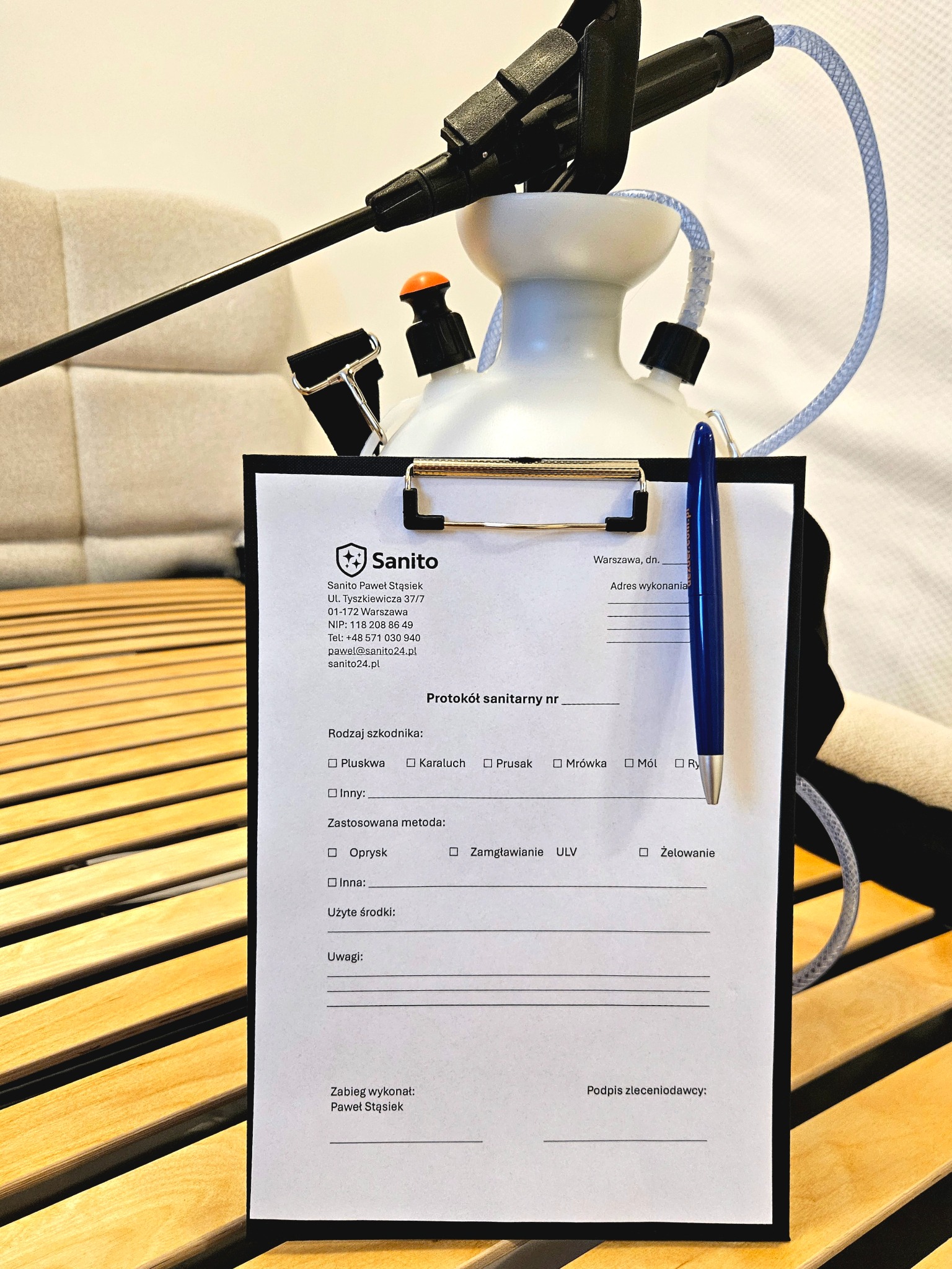Opryskiwacz z protokołem sanitarnym Sanito na drewnianym stelażu łóżka. Protokół z zaznaczonymi polami wyboru szkodnika i metodą oprysku. Niebieski długopis.