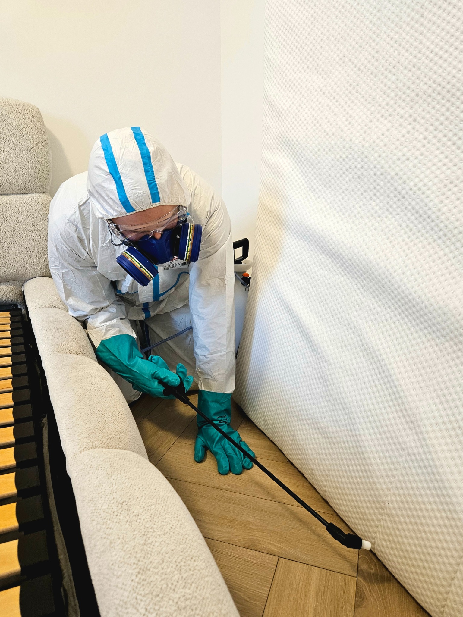 Pracownik w kombinezonie ochronnym, masce i rękawicach aplikuje preparat za pomocą lancy na materac łóżka. Zabieg wykonywany w pomieszczeniu.