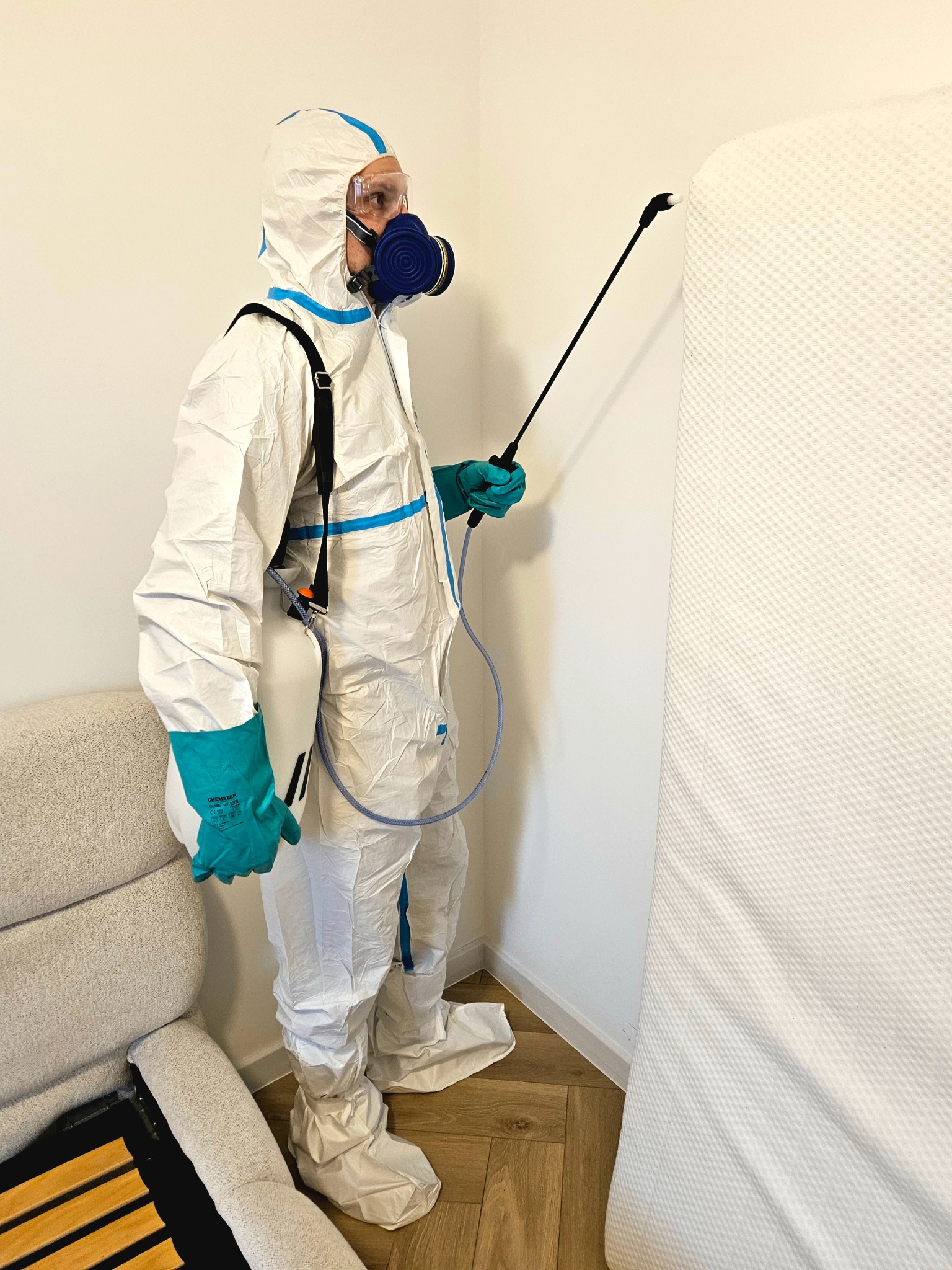 Osoba w kombinezonie ochronnym i masce spryskuje materac. Widoczny aplikator, butla na plecach i fragment sofy. Dezynfekcja materaca w pomieszczeniu.