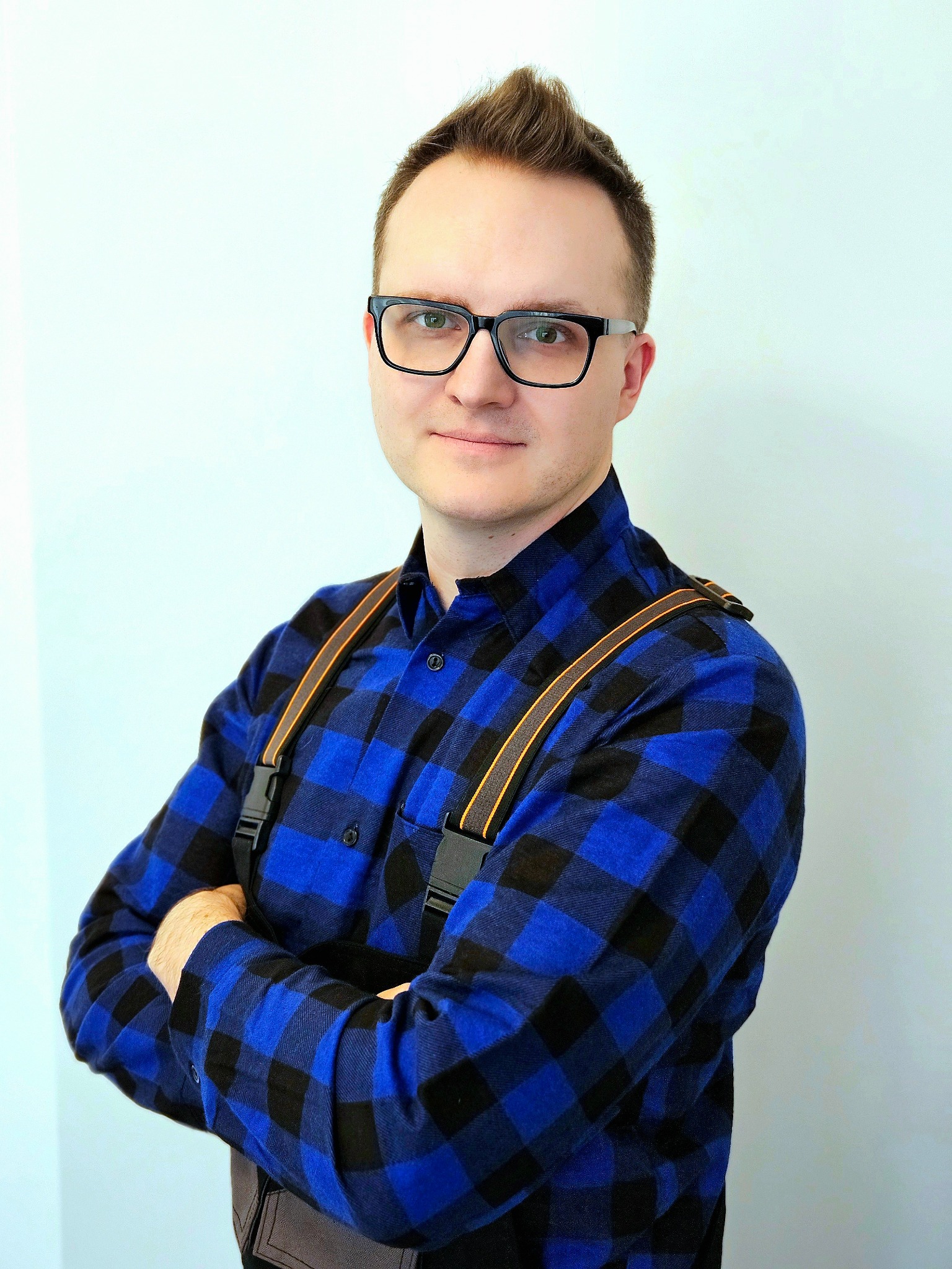 Portret mężczyzny w okularach i niebieskiej koszuli w kratę, z założonymi rękami, na jasnym tle. Widoczne szelki i fragment pasa narzędziowego.