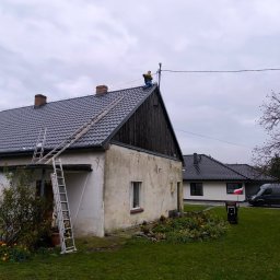 Mikołaj Długowski - Praca na dachu: Elektryk w żółtej kurtce montuje instalację odgromową na szczycie domu z ciemną dachówką. Dwie drabiny opierają się o ścianę. Pochmurne niebo w tle.