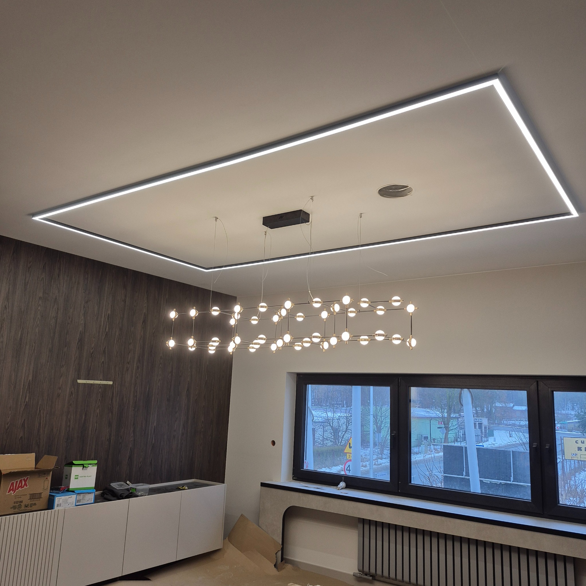 Nowoczesne oświetlenie sufitowe: prostokątna listwa LED i designerska lampa wisząca z kulistymi żarówkami w jasnym wnętrzu. Widok na okno i fragment mebli.