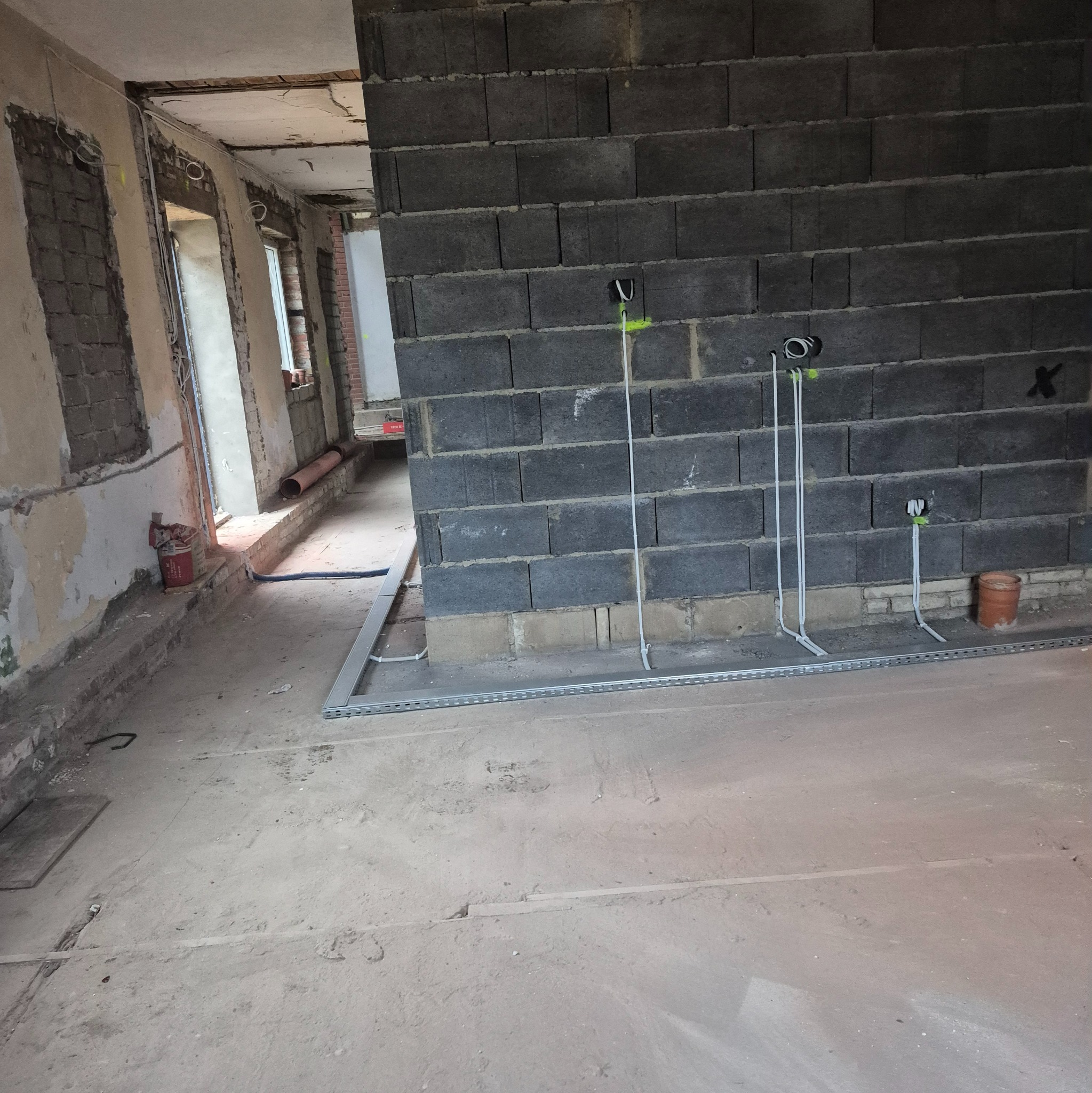 Surowy stan wnętrza w trakcie prac: widoczne przewody elektryczne wyprowadzone ze ściany z bloczków betonowych, koryto kablowe na podłodze, ściany w stanie deweloperskim.