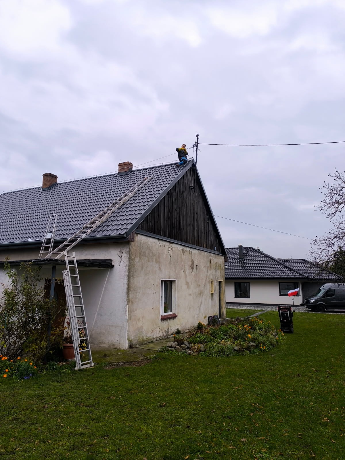 Praca na dachu: Elektryk w żółtej kurtce montuje instalację odgromową na szczycie domu z ciemną dachówką. Dwie drabiny opierają się o ścianę. Pochmurne niebo w tle.