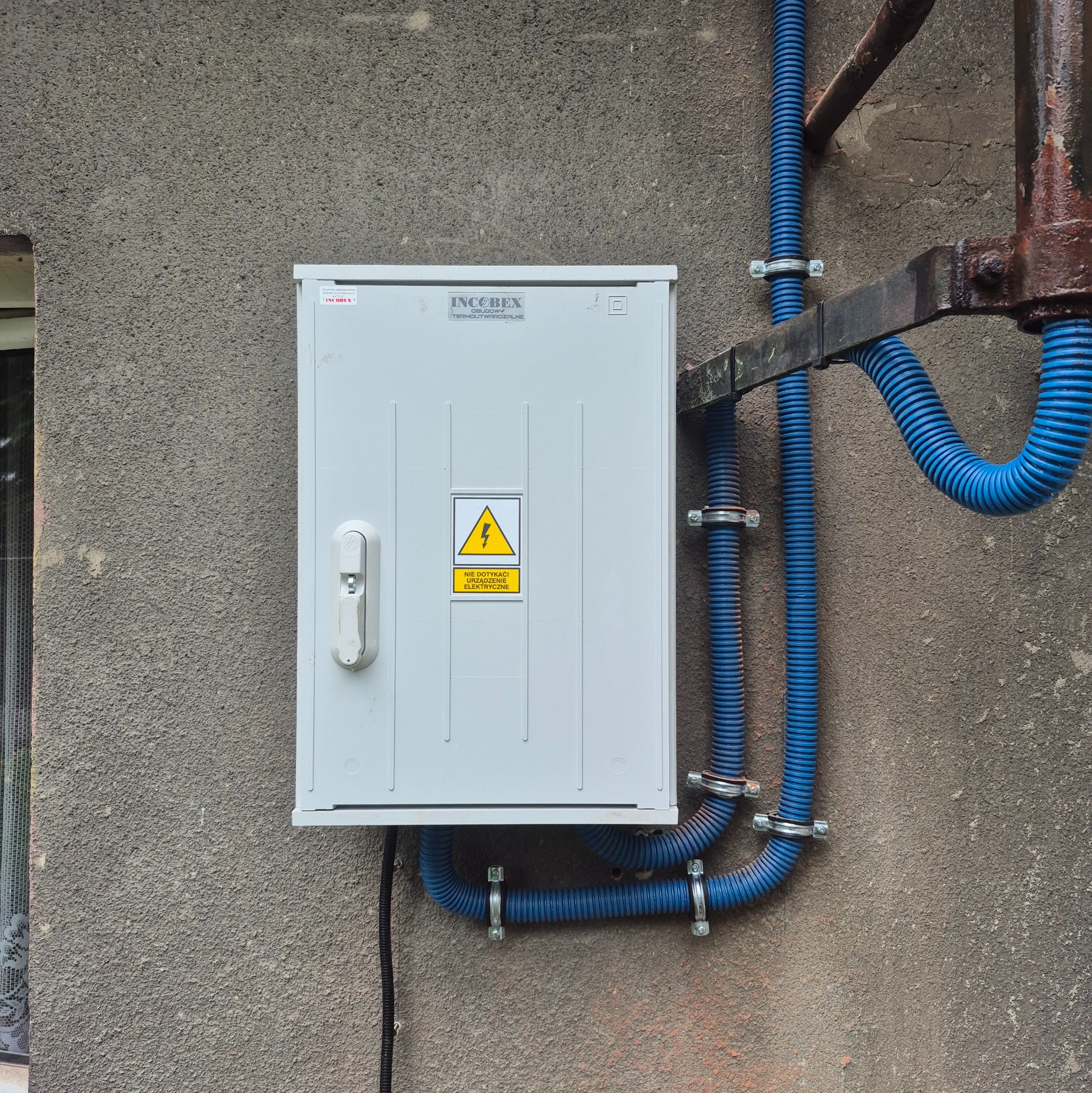 Biała skrzynka elektryczna z ostrzeżeniem o napięciu, zamontowana na szarej, chropowatej ścianie. Niebieskie rury ochronne prowadzą do skrzynki, przymocowane metalowymi obejmami.