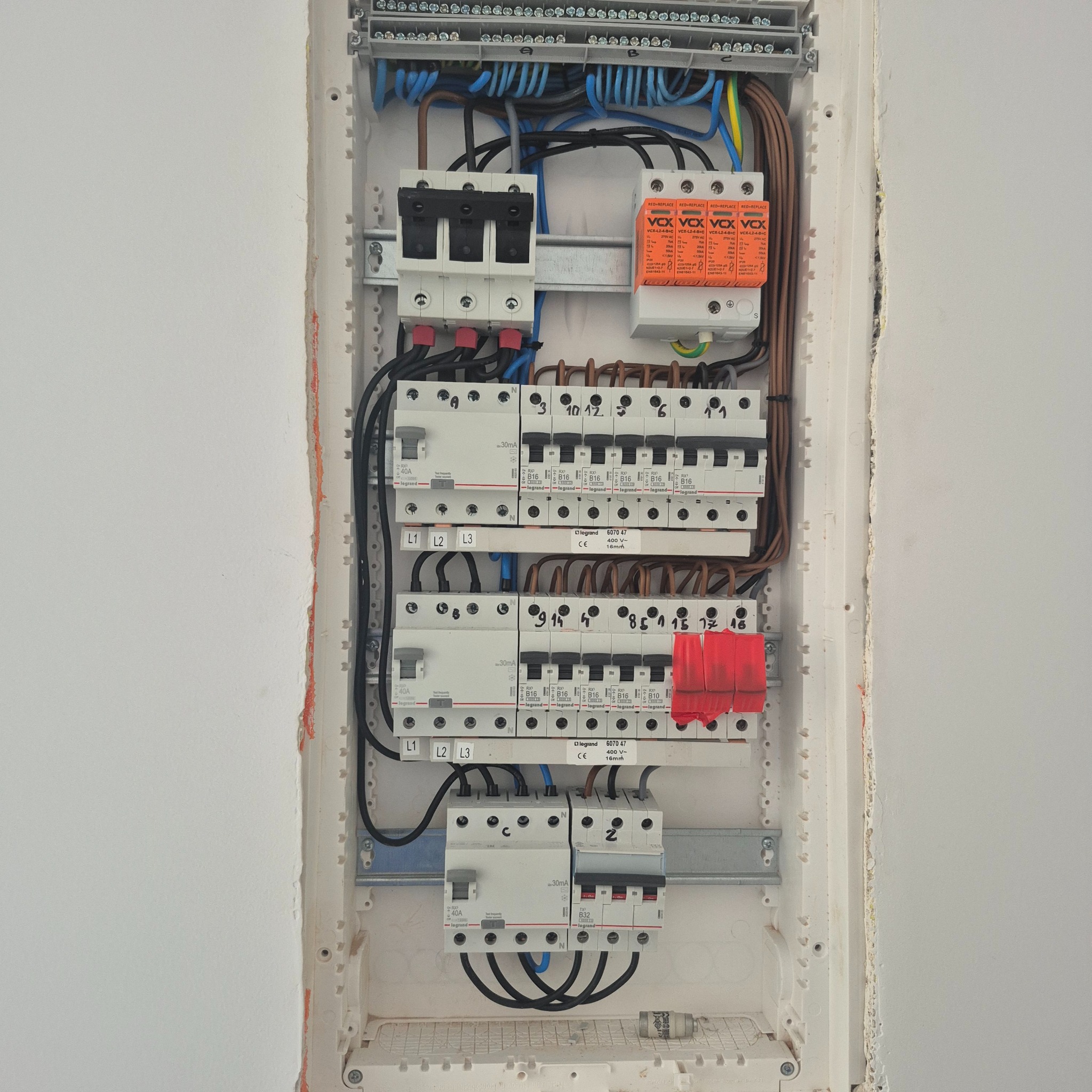 Rozdzielnica elektryczna z zainstalowanymi bezpiecznikami i przewodami wewnątrz białej obudowy, widok z bliska na ścianie. Elementy oznaczone numerami i symbolami.