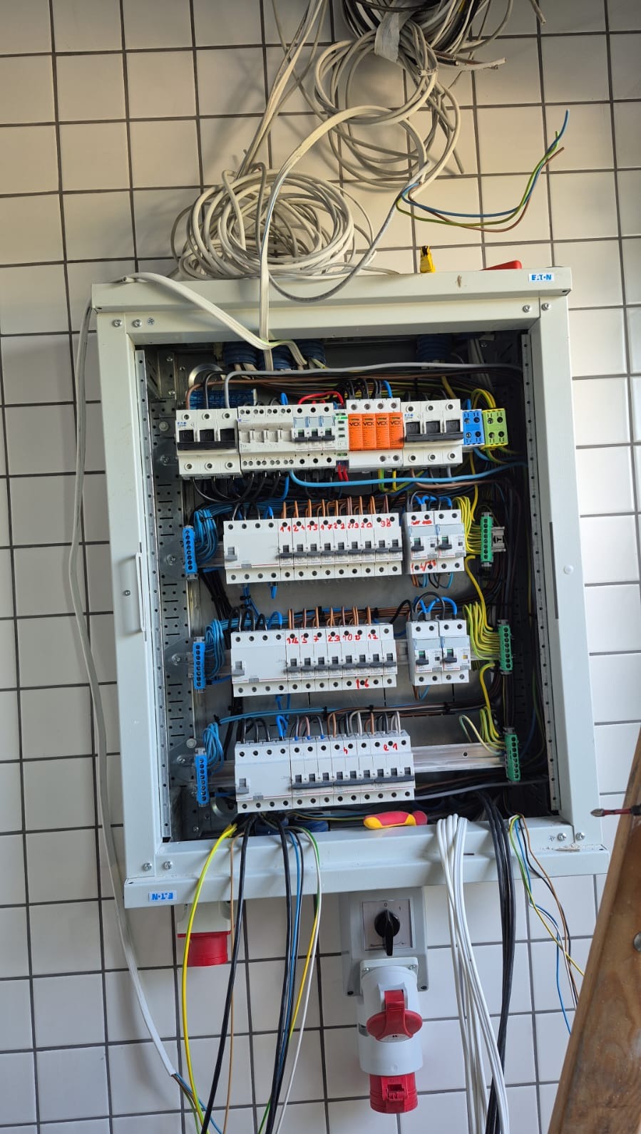 Otwarta skrzynka elektryczna z bezpiecznikami i przewodami na kafelkowanej ścianie. Widoczne gniazda siłowe i część drabiny. Przewody ułożone wewnątrz i na zewnątrz skrzynki.