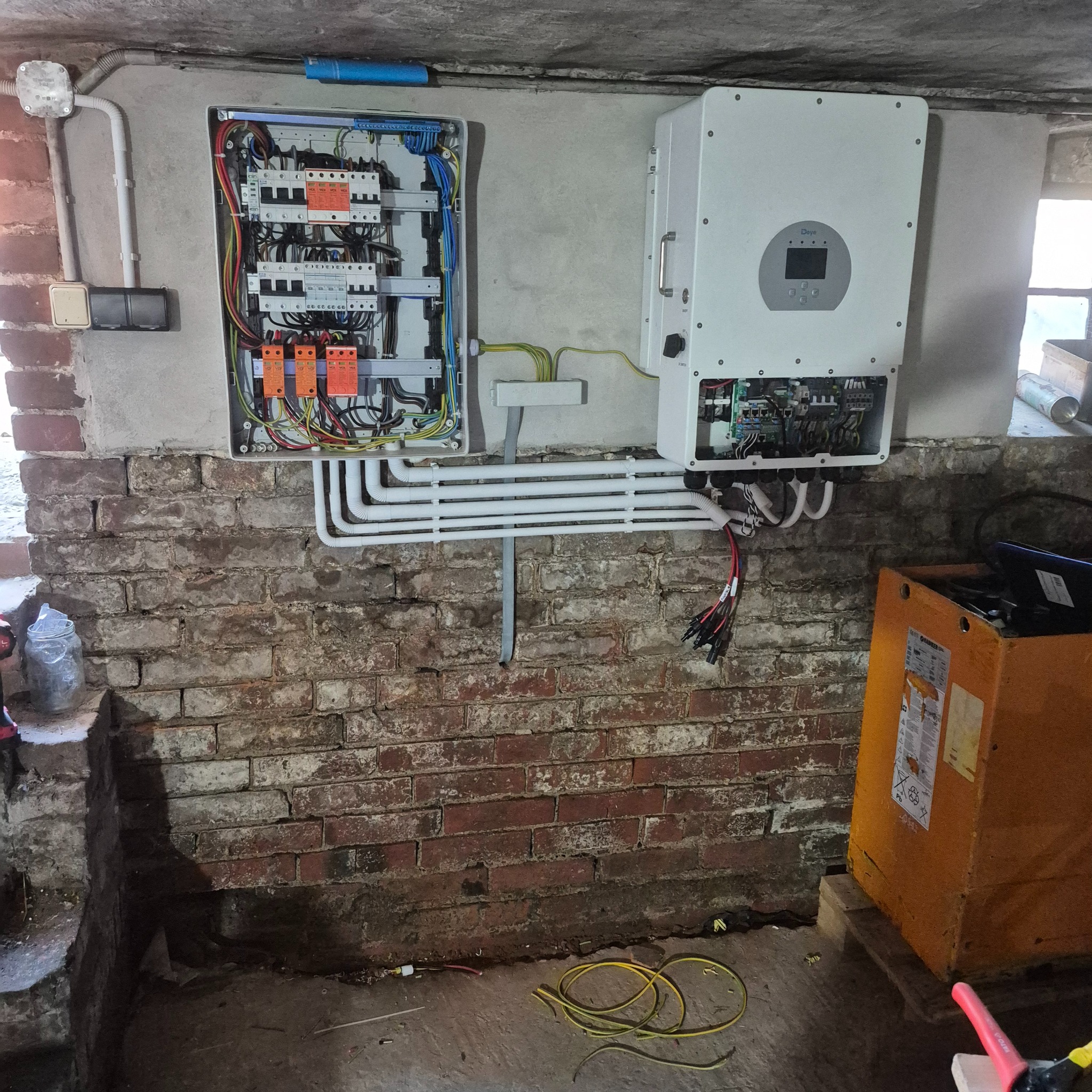 Rozdzielnia elektryczna z falownikiem na ceglanej ścianie piwnicy. Widoczne kable i przewody. Instalacja w trakcie montażu, skrzynka otwarta.
