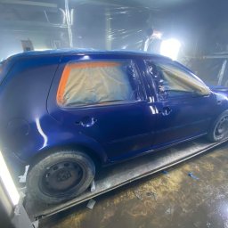 Paint Garage - Niebieski samochód w kabinie lakierniczej, częściowo oklejony taśmą i folią, przygotowany do lakierowania. Widoczne oświetlenie i podłoga kabiny.
