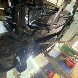 Paint Garage - Podwozie samochodu po konserwacji antykorozyjnej, uniesione na podnośniku w warsztacie. Widoczne narzędzia i oświetlenie warsztatowe.