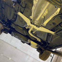 Paint Garage - Podwozie samochodu na podnośniku po konserwacji antykorozyjnej, widoczne elementy zawieszenia i układu wydechowego pokryte ochronną powłoką. Detal: oświetlenie warsztatowe.
