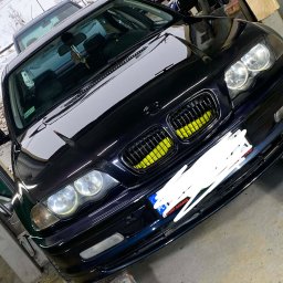 Paint Garage - Błyszczący, czarny BMW E46 po tuningu grilla na żółto, zaparkowany w warsztacie. Widoczne detale karoserii i reflektory. W tle inne pojazdy i zimowy krajobraz.