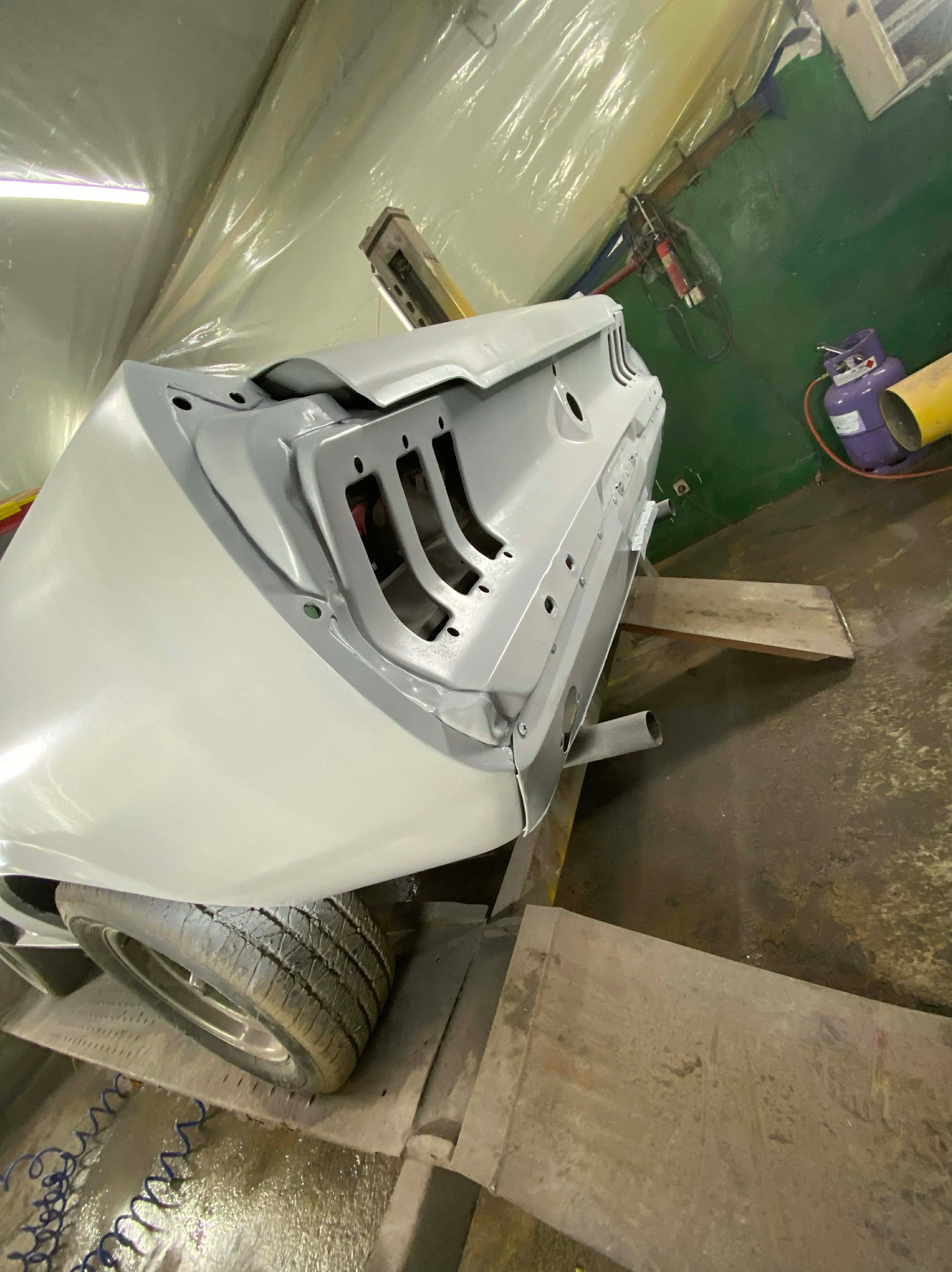 Szary, odrestaurowany tył klasycznego Mustanga w warsztacie, widoczny fragment opony, podnośnik i butla z gazem. Świeżo położony podkład lakierniczy.