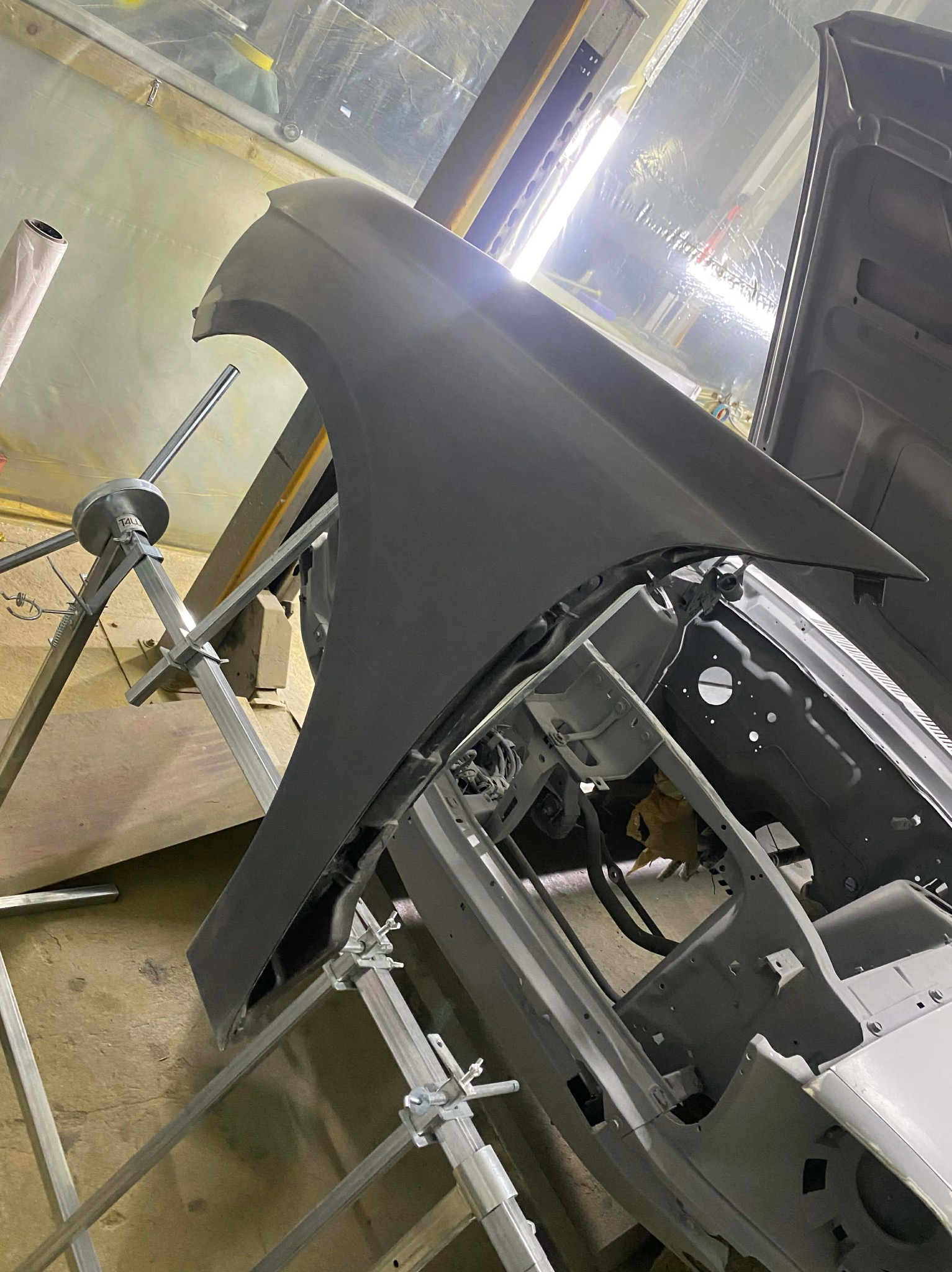 Elementy karoserii auta w trakcie przygotowania do lakierowania, umieszczone na metalowym stelażu w warsztacie. Widoczny fragment maski i błotnik.