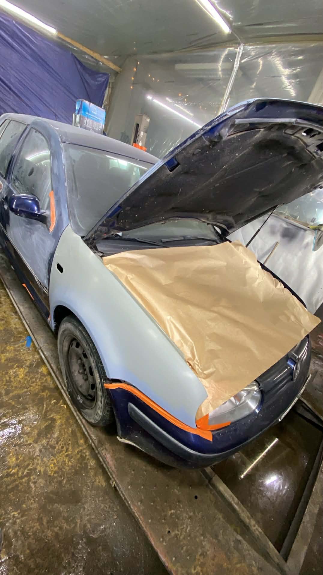 Niebieski samochód w warsztacie z otwartą maską, przygotowany do lakierowania. Nowy, szary błotnik i papier maskujący na masce. Taśma malarska zabezpiecza elementy.