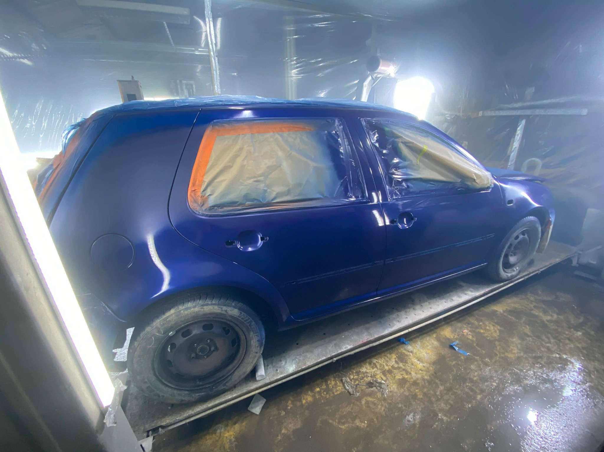 Niebieski samochód w kabinie lakierniczej, częściowo oklejony taśmą i folią, przygotowany do lakierowania. Widoczne oświetlenie i podłoga kabiny.