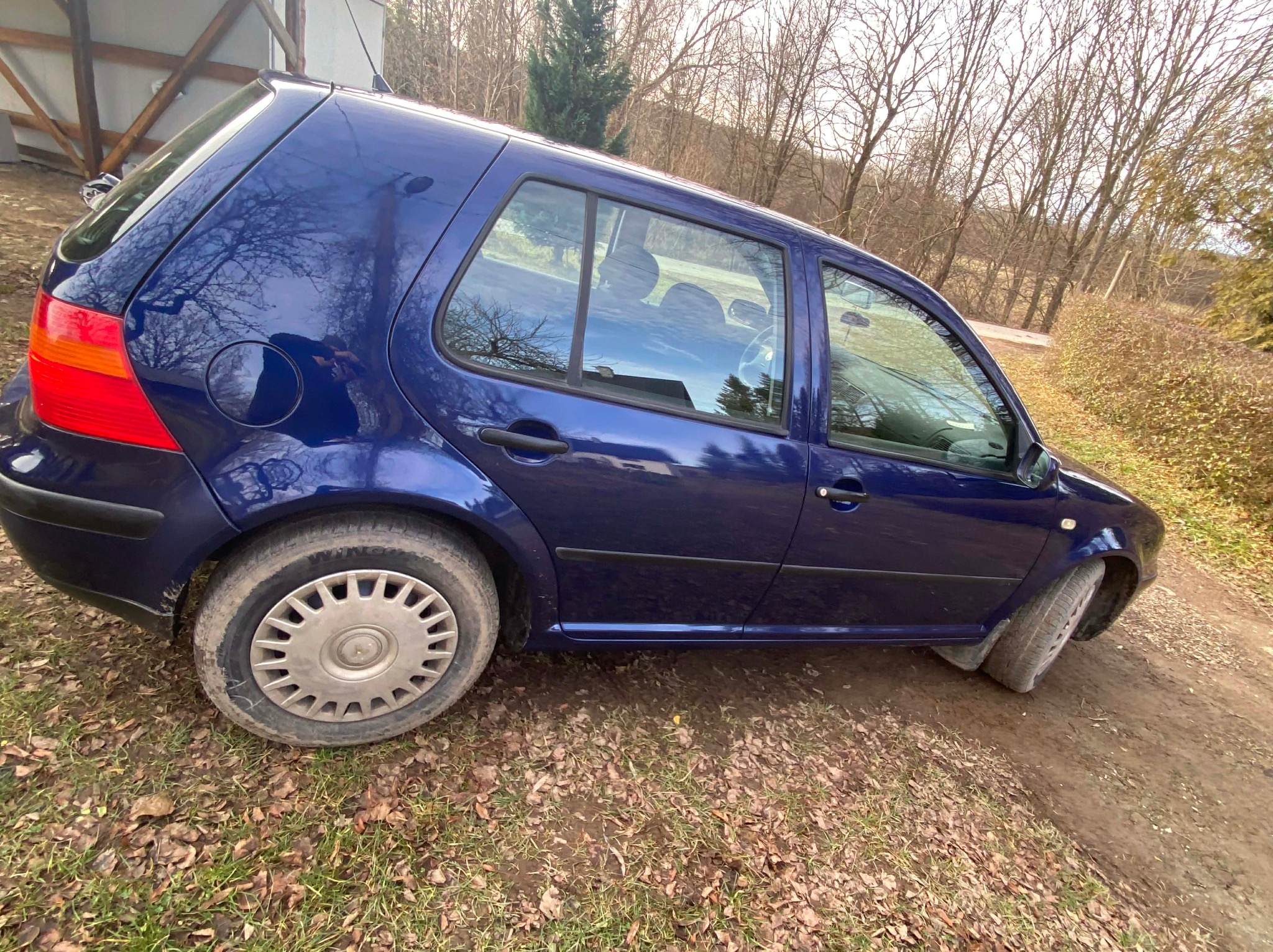 Niebieski hatchback na polnej drodze, widok z boku. Auto z lekko zabrudzonymi oponami, tło stanowią drzewa bez liści i fragment budynku.