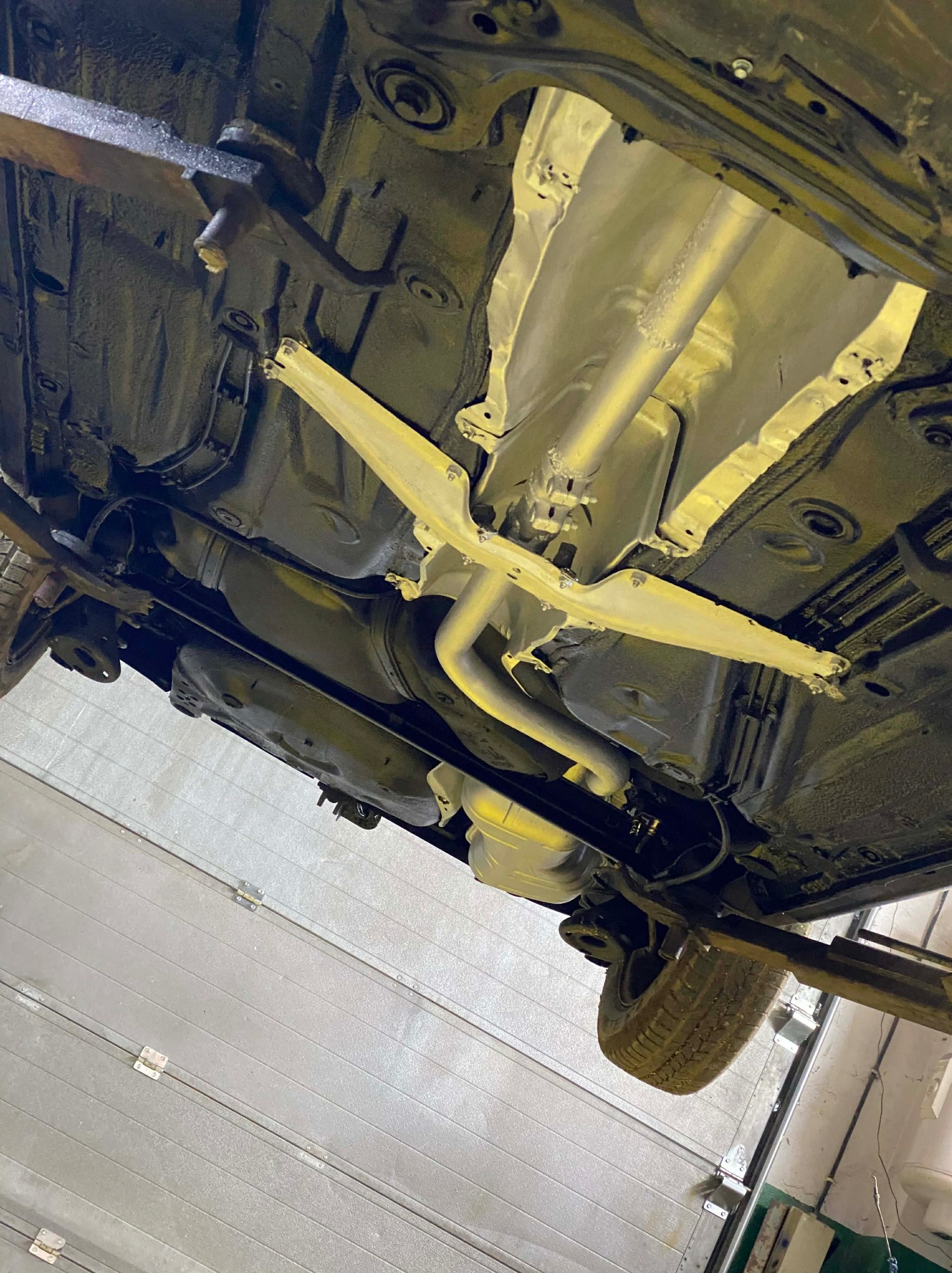 Podwozie samochodu na podnośniku po konserwacji antykorozyjnej, widoczne elementy zawieszenia i układu wydechowego pokryte ochronną powłoką. Detal: oświetlenie warsztatowe.