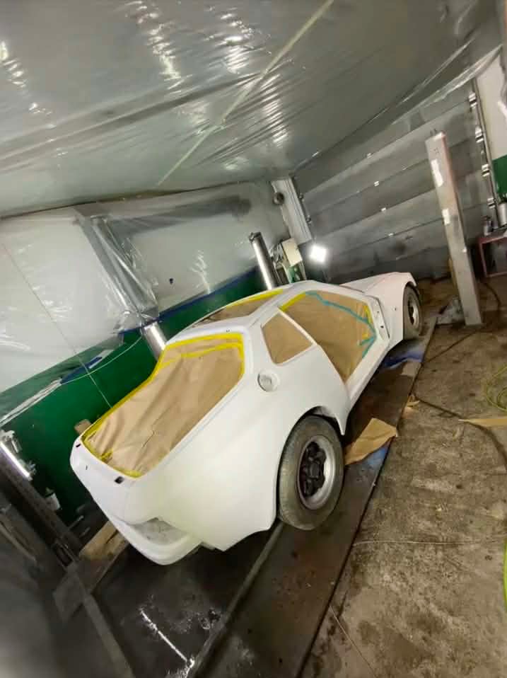 Biały samochód sportowy, częściowo oklejony taśmą malarską i papierem, przygotowany do lakierowania w warsztacie. Widoczne detale karoserii i opony.