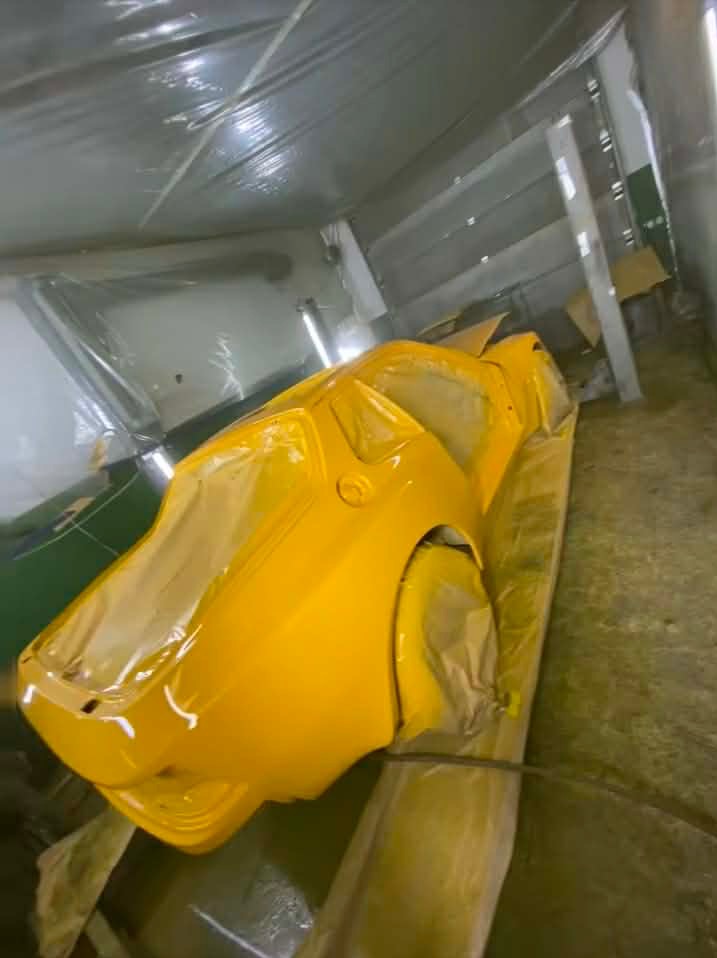 Żółty samochód sportowy w trakcie lakierowania w kabinie lakierniczej, częściowo zakryty folią ochronną. Widoczne detale karoserii i oświetlenie warsztatowe.