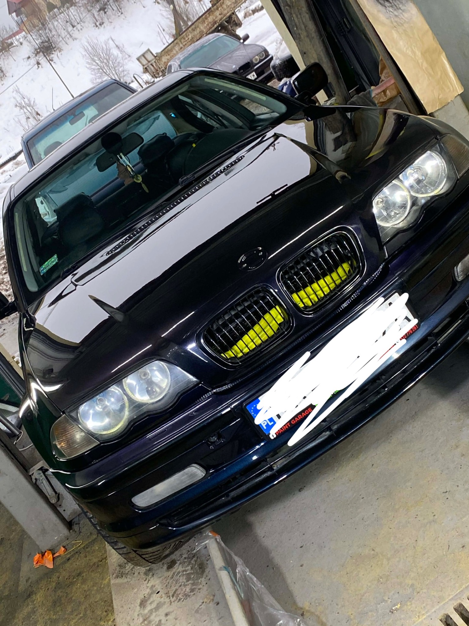 Błyszczący, czarny BMW E46 po tuningu grilla na żółto, zaparkowany w warsztacie. Widoczne detale karoserii i reflektory. W tle inne pojazdy i zimowy krajobraz.