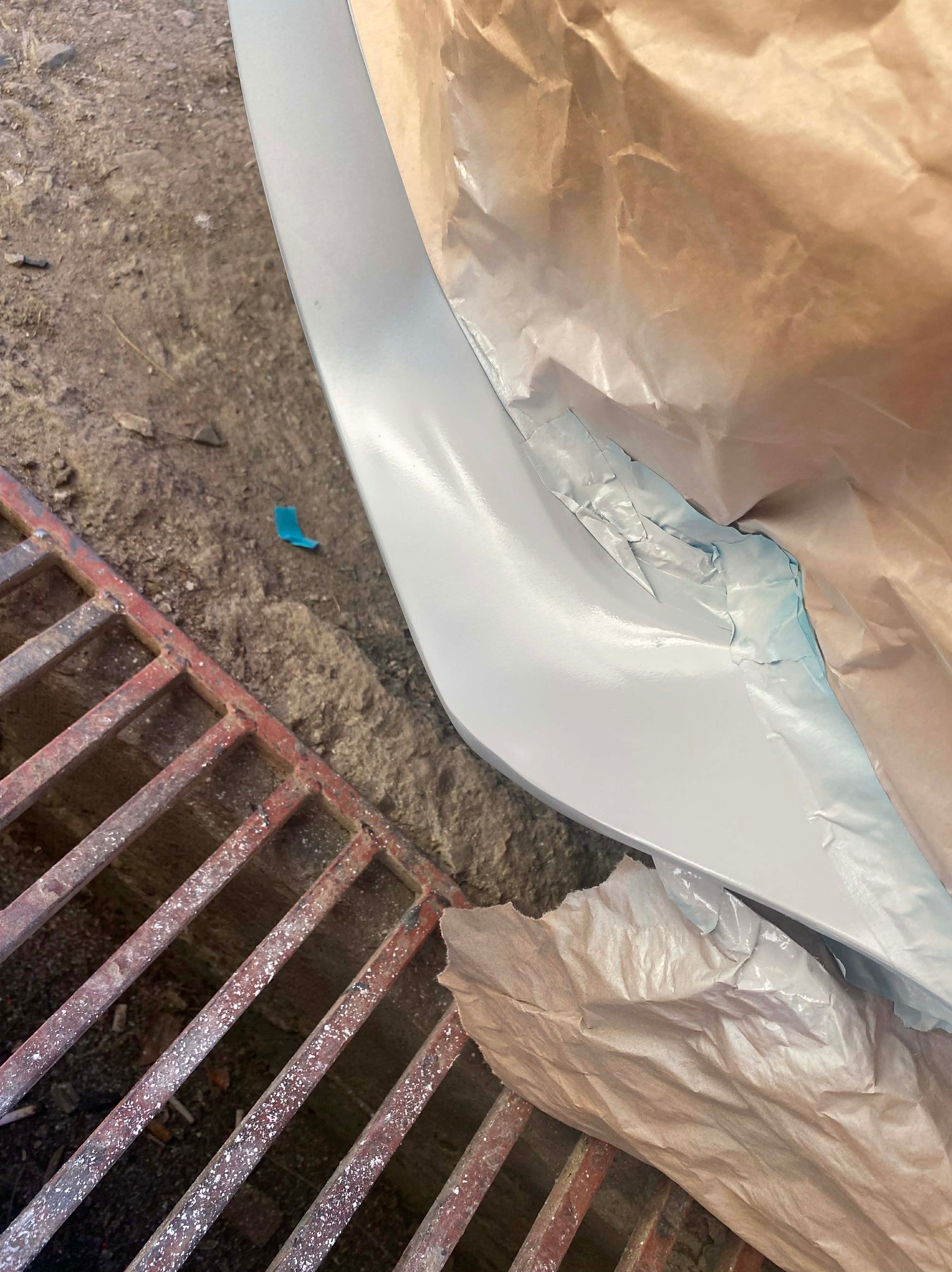 Pomalowany na szaro element karoserii samochodowej, zabezpieczony papierem i taśmą, leży na betonowej posadzce obok metalowej kratki ściekowej.
