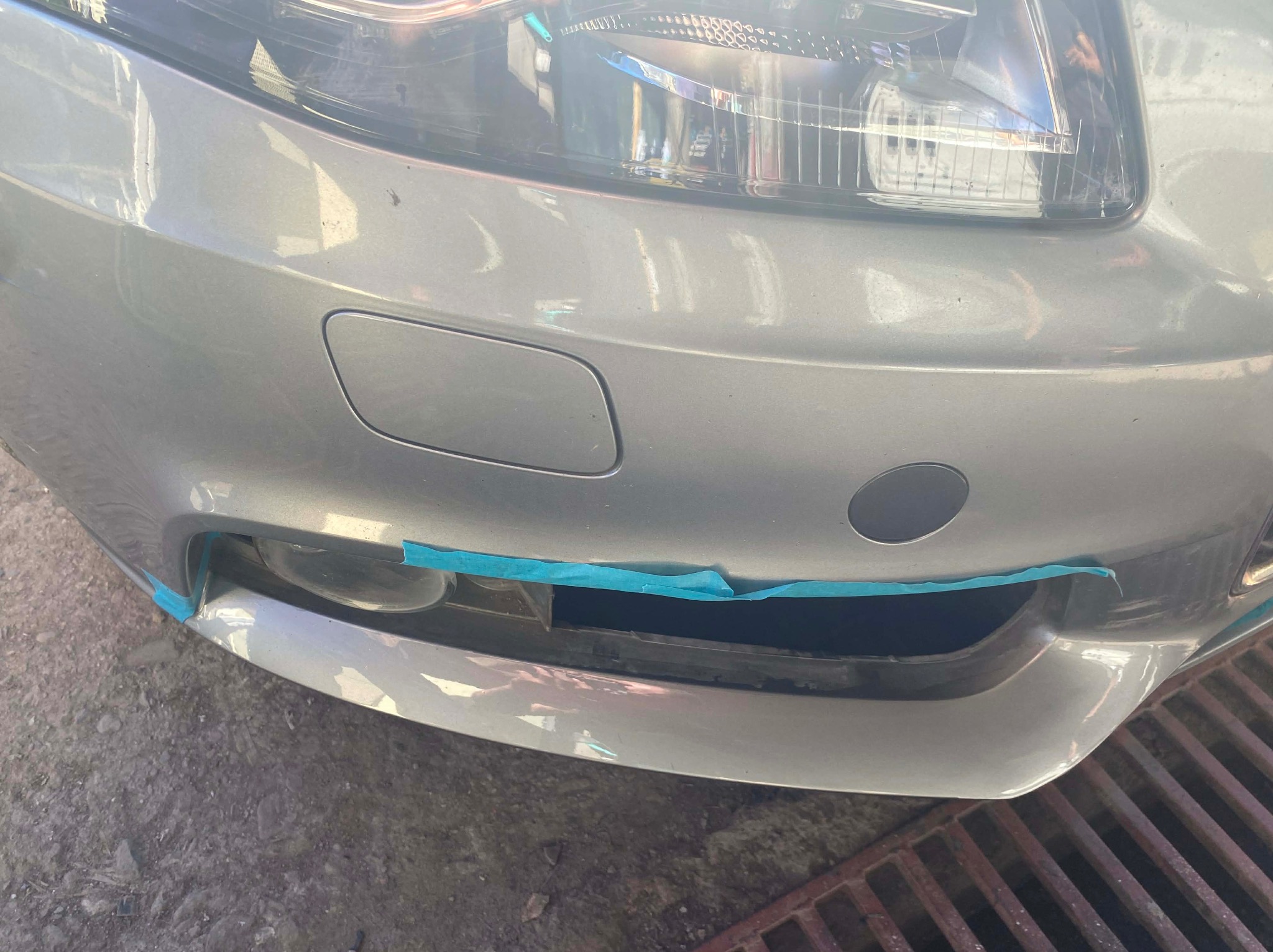 Szary samochód z niebieską taśmą maskującą na przednim zderzaku, przygotowany do naprawy lub lakierowania. Widoczny fragment kratki ściekowej w tle.