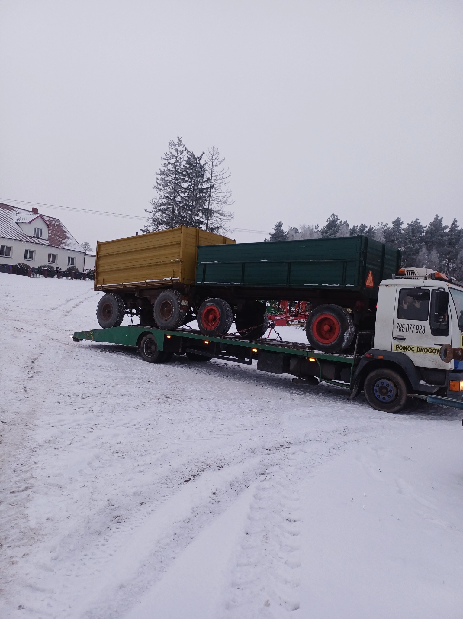 Laweta przewozi dwa traktory z przyczepami w zimowej scenerii. Na białym samochodzie napis 'Pomoc Drogowa' i numer telefonu. W tle dom i drzewa.