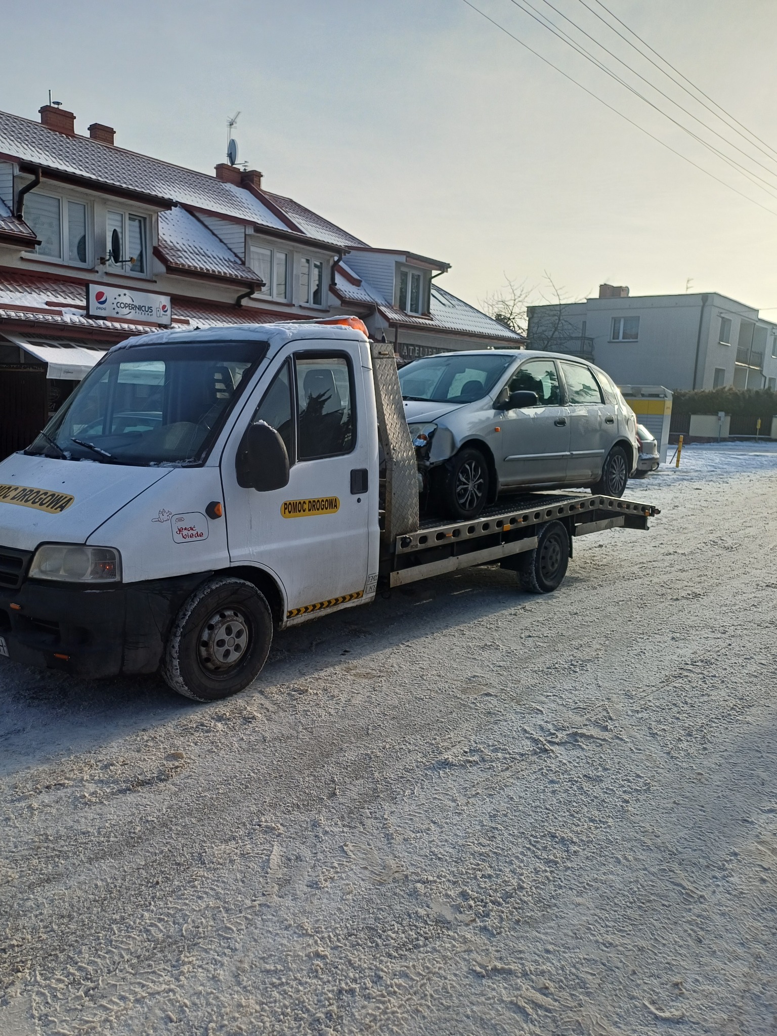 Laweta z uszkodzonym srebrnym autem na zaśnieżonej drodze. Na aucie naklejka 'Pomoc Drogowa'. W tle budynki mieszkalne i szyld 'Copernicus'.