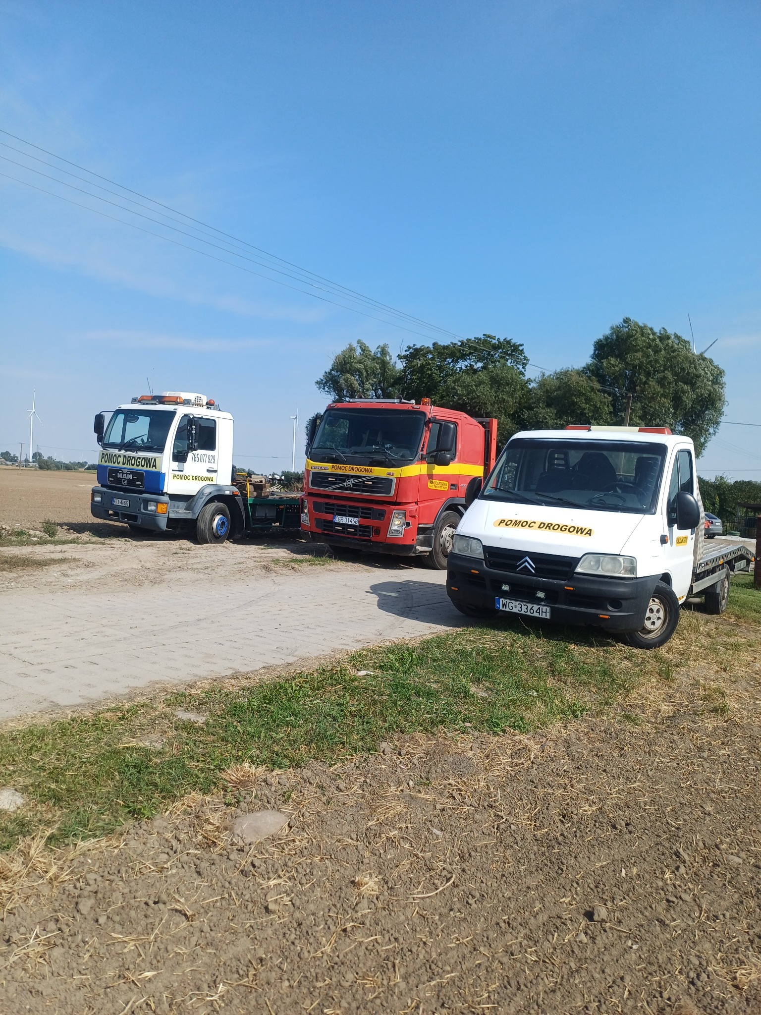 Trzy pojazdy pomocy drogowej (MAN, Volvo, Citroen) na poboczu w Radzyniu Chełmińskim. W tle widać turbiny wiatrowe. Słoneczny dzień, błękitne niebo.