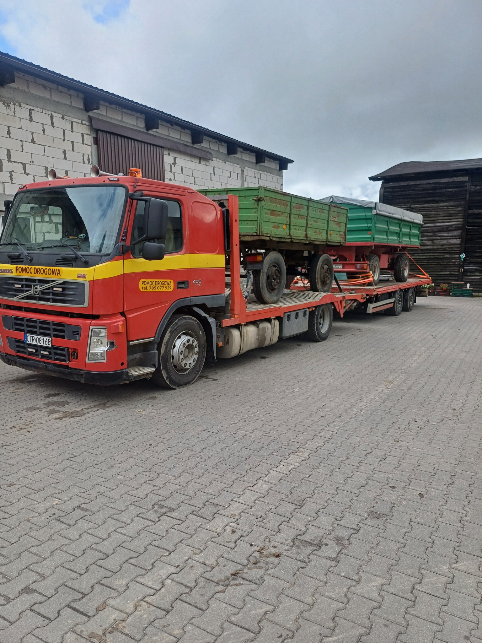 Czerwona laweta Volvo z żółtym pasem i napisem 'Pomoc Drogowa' przewozi dwie zielone przyczepy rolnicze na tle szarej kostki brukowej i budynków gospodarczych.
