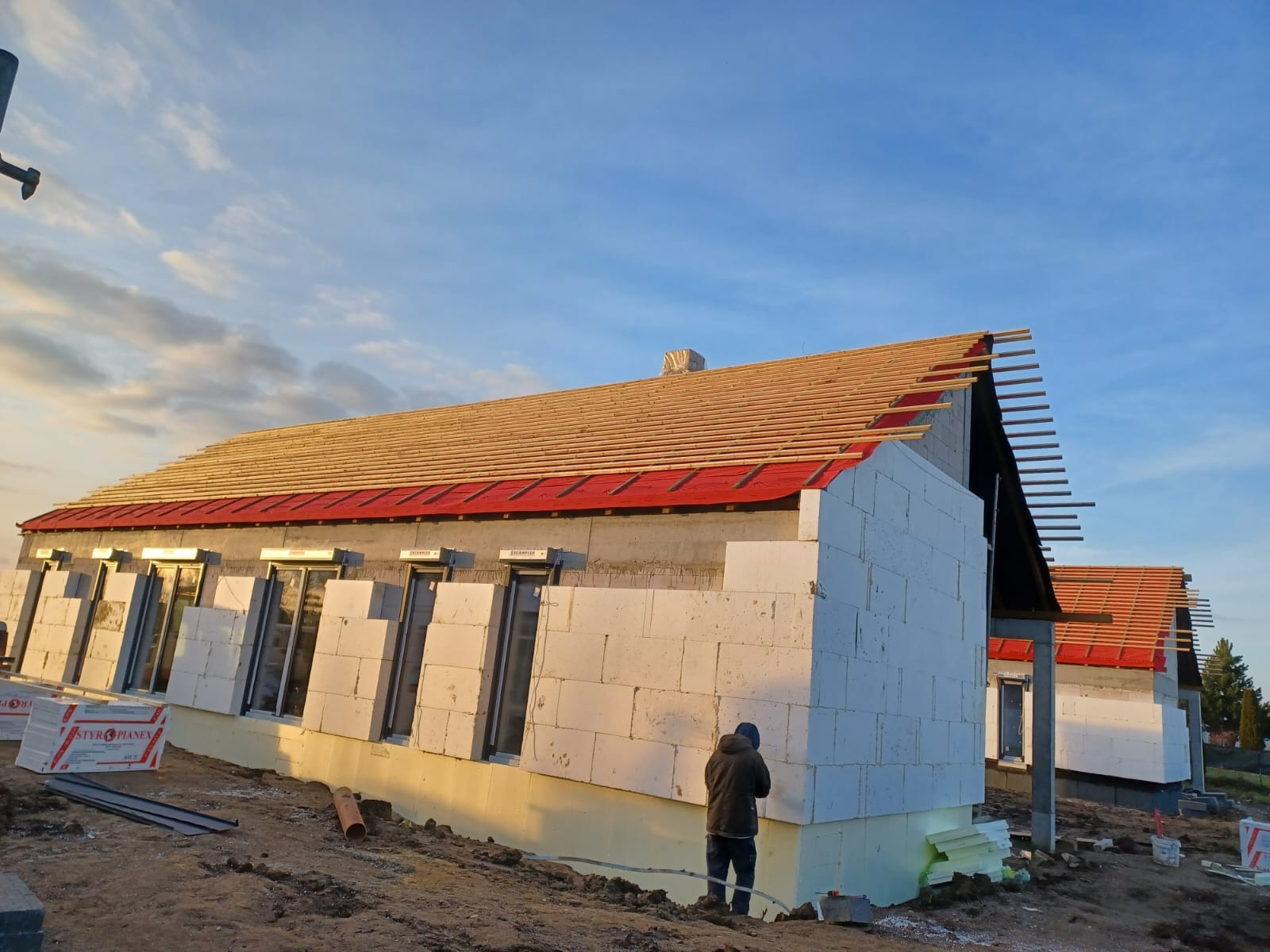 Budowa domu jednorodzinnego: widok na konstrukcję dachu z łatami na czerwonej membranie, ściany z białego betonu komórkowego, okna wstawione, ocieplenie fundamentów.