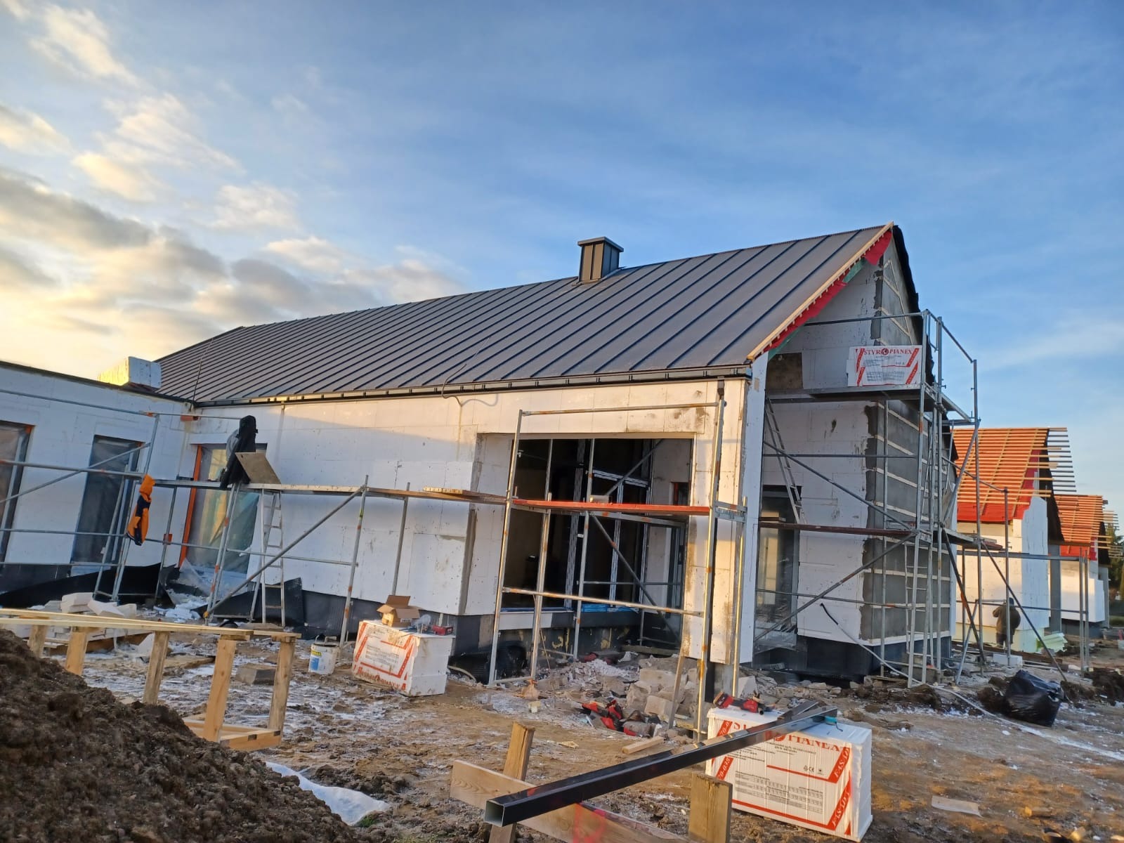 Budowa domu jednorodzinnego z szarą blachodachówką, ocieplonego styropianem, z widocznymi rusztowaniami i elementami konstrukcyjnymi w słoneczny dzień.