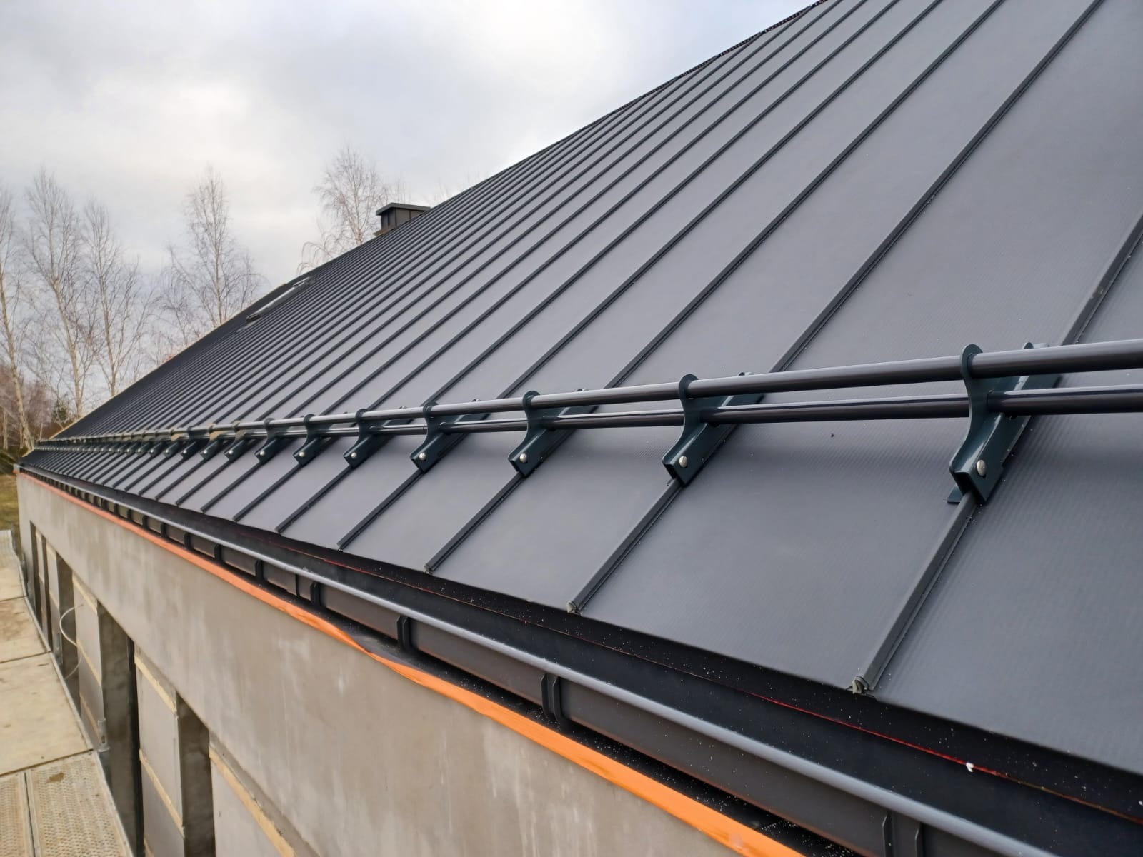 Grafitowy dach z blachy na rąbek stojący z systemem przeciwśniegowym, widoczny z dołu pod kątem. Szara elewacja budynku w tle, pochmurne niebo.