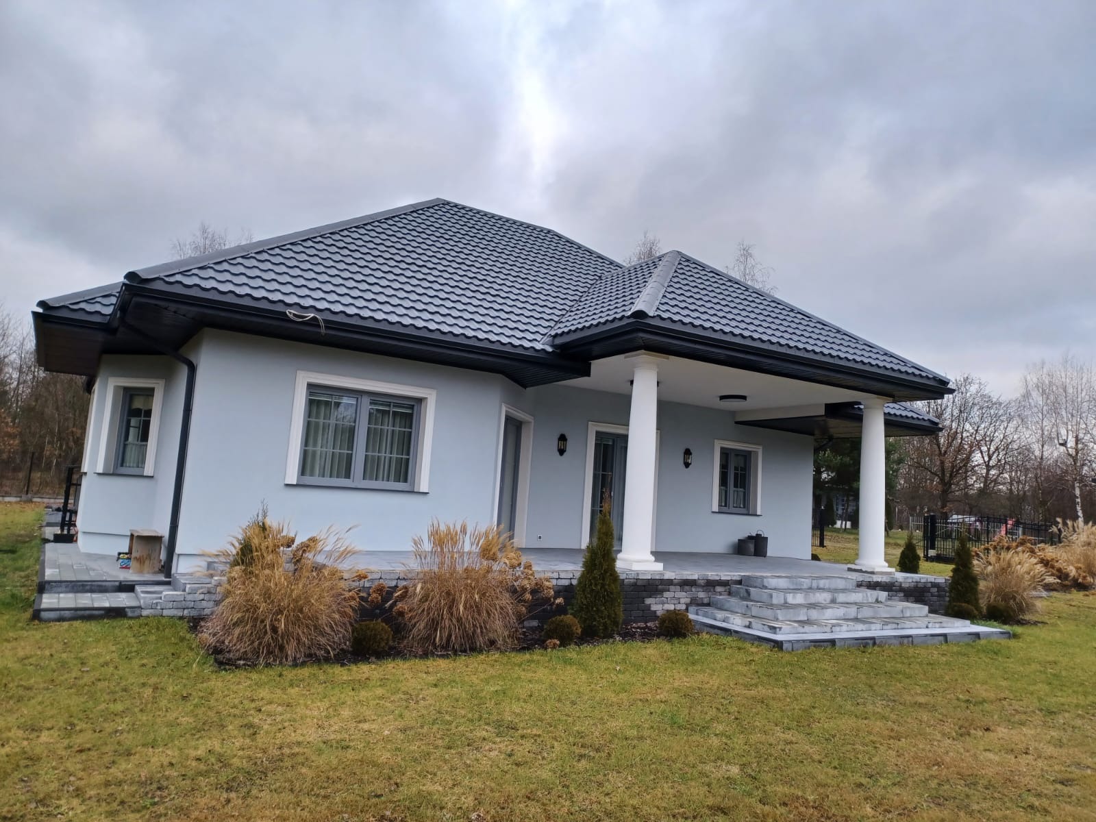 Dom jednorodzinny z ciemnoszarą dachówką karpiówką i czarnymi rynnami, widok z ogrodu w pochmurny dzień. Elewacja w odcieniach szarości, białe kolumny przy wejściu.