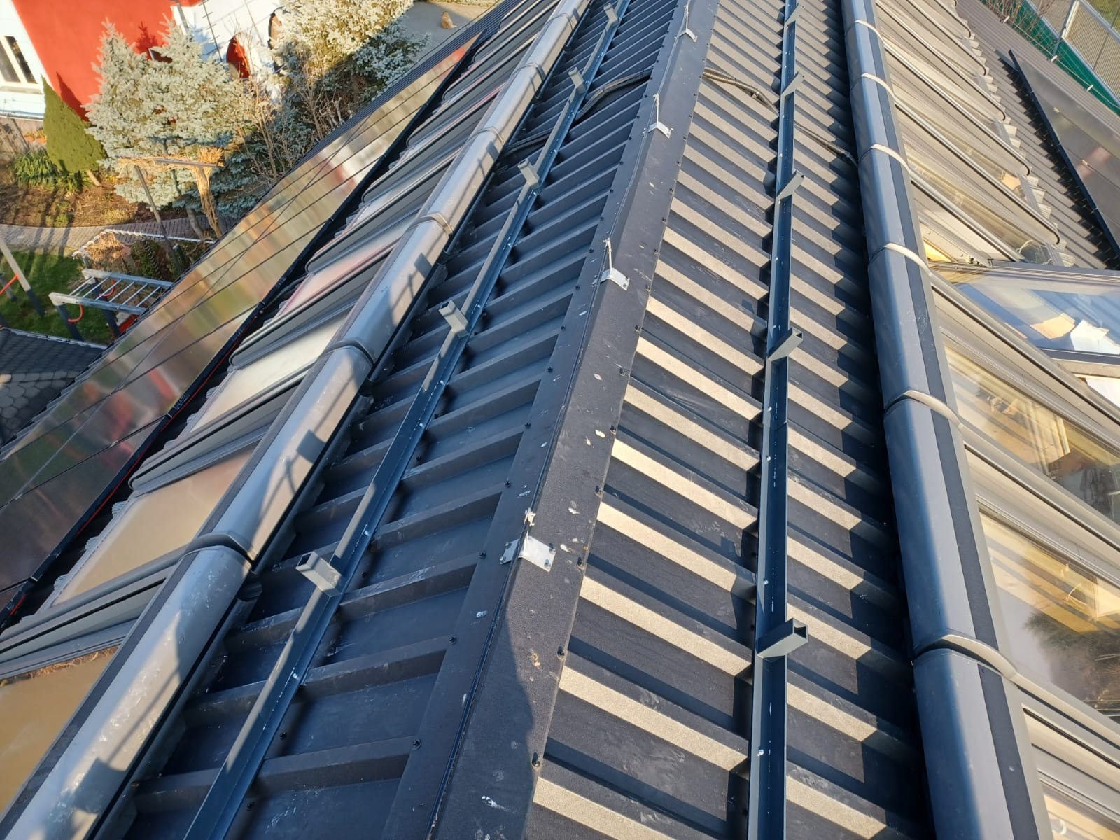 Czarny dach z blachy trapezowej z oknami dachowymi i elementami orynnowania, widok z góry w słoneczny dzień. Charakterystyczne mocowania rynien.