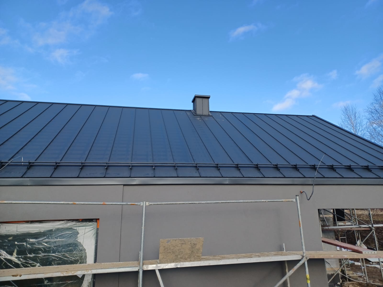 Nowoczesny, grafitowy dach z blachy na szarym budynku, widoczny komin i rusztowania. Błękitne niebo w tle. Charakterystyczne mocowania rynien.