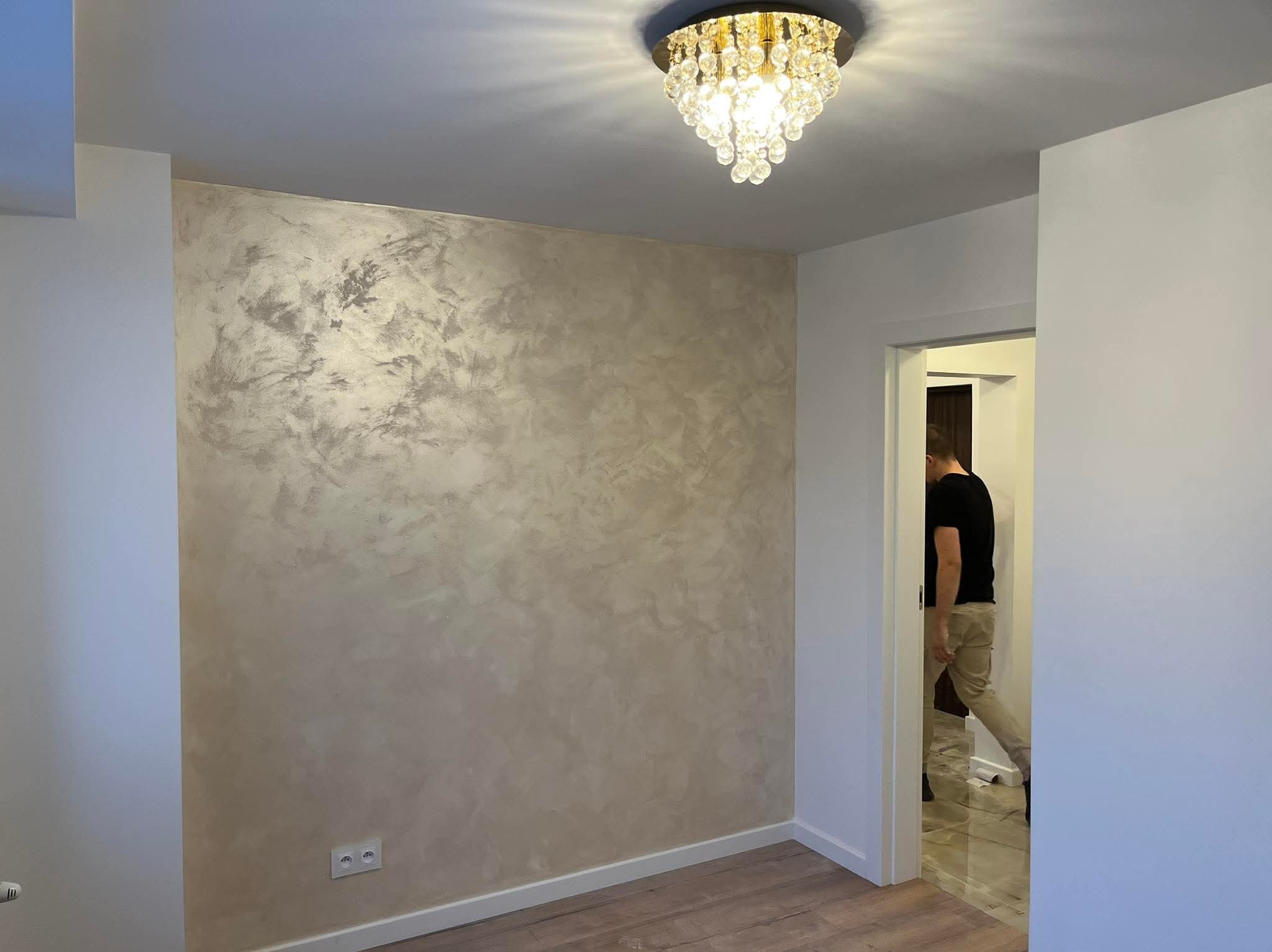 Ściana z dekoracyjnym efektem stiuku w odcieniach beżu i srebra, oświetlona kryształowym żyrandolem. Widoczny fragment podłogi z paneli i osoba w przejściu.